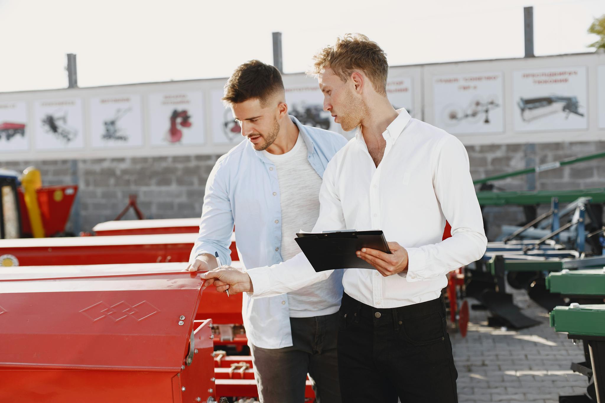 Two men discussing agricultural equipment purchase at a dealership, one holding a clipboard outside.